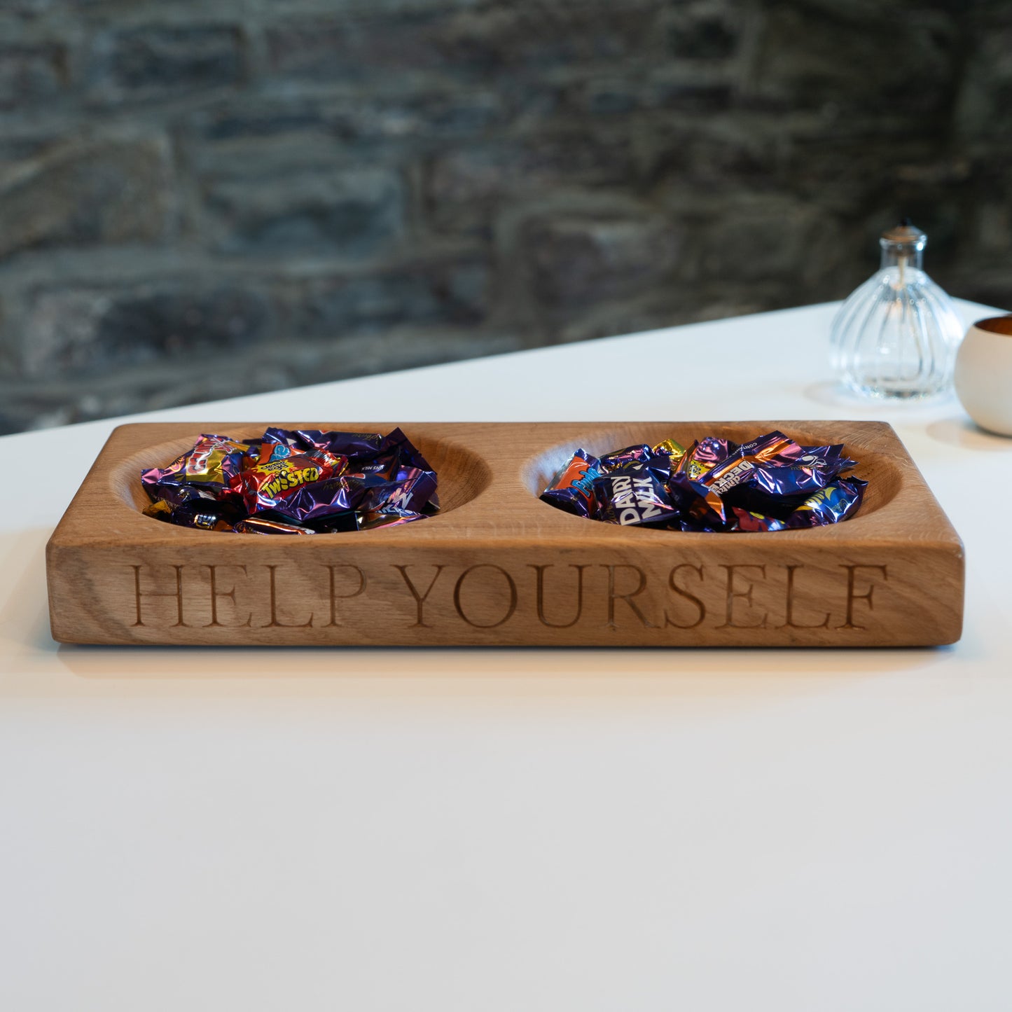 Wooden tray with two compartments filled with chocolates, engraved with 'Help Yourself', on a white surface with a stone wall background.
