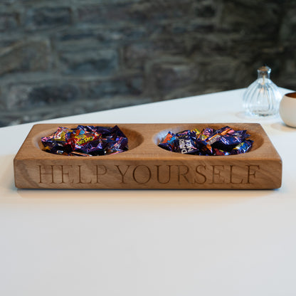 Wooden tray with two compartments filled with chocolates, engraved with 'Help Yourself', on a white surface with a stone wall background.